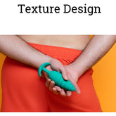 A person in red shorts holds the B-Vibe Texture Vibrating Butt Plug Twist - Green (Large) behind their back. The orange background features "Texture Design" text above. The product, by B-Vibe, is made of body-safe silicone and has a textured, vibrating design.